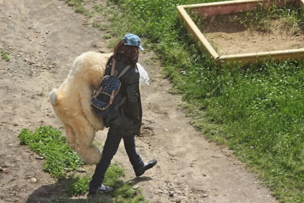 A tramp woman in a black leather jacket carries a stuffed toy a large plush Bunny on a summer day behind her back on sand box background in Park