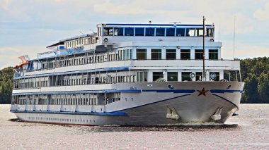 Güzel Rusya seyahati. Dört güverte beyaz yolcu gemisi. Nehirde yüzen yolcu gemisi. Turist gemisi, yaz günü nehir kenarında yeşil ağaçlar ve mavi gökyüzü arka planıyla kapanıyor.