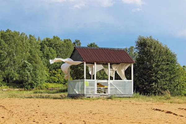 Abandoned lonely gazebo with curtains fluttering in the wind on a deserted sandy beach against the background of a green forest on a Sunny summer day, windy weather, storm