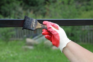 Beyaz kırmızı iş eldivenli bir el, fırçayla boyar. Yatay metal bir çit. Bir yaz günü, bir köyde siyah ziftle boyanmış.