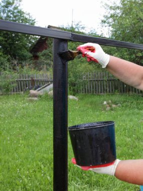 Kaynaklı eklem yerlerinin kaynamış tablosu. Eldivenin içindeki bir el, kaynaklı çit destek direğini boyuyor. Yazın yeşil çimlerin arka planına siyah bituminöz boya ile kaplıyor.
