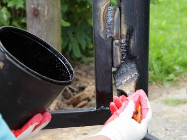 Bir iş eldiveni içindeki bir el, kaynaklı kapı menteşesini fırçayla boyar, arka plandaki boya kovasının arka planına yapıştırır. Kaynak işçiliğinin önleyici tablosu.