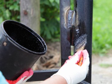 Anticorrosive painting of welded joints, a hand in a white red work glove paints the welded gate door hinge with black bituminous paint against the background of a plastic paint can close up clipart