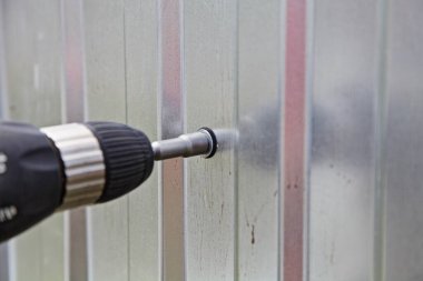 Endüstriyel işler, soket anahtarı olan elektrikli matkap tornavidayla Chuck vidaları sökerek yeni gri metal profilli bir levhaya, çinko kaplı bir çit inşa ediyor.
