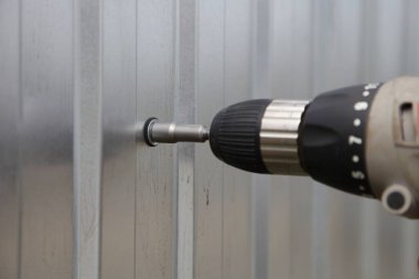 Endüstriyel bağlama, vidaları yeni gri metal bir levhaya vidalamak için kısa devre anahtarı takılı elektrikli matkap tornavidası, çinko kaplı bir çit inşaatı.