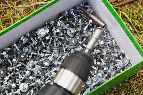 A lot of new gray galvanized metal screws in a box on green grass against the background of a blurred quick-release screw driver cartridge