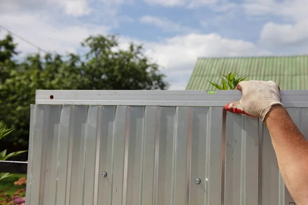 Construction of an metal profiled sheet fence, a Man\'s hand checks with level tool its horizontal mounting on a summer Sunny day