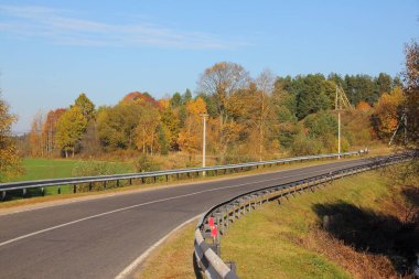 Boş kuru asfaltlanmış kır yolu güvenli metal çitlerle donatılmış yeşil sarı ağaçlar ve mavi gökyüzü arka planı güneşli ekim gününde, güzel kırsal alan, güz arabasının güvenliği
