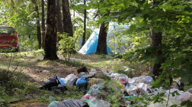 Güneşli bir yaz gününde ormanda kamp çadırının ve arabanın yanında büyük bir çöp yığını. Çevreyi turistlerle dolduran sosyal sorunlar.