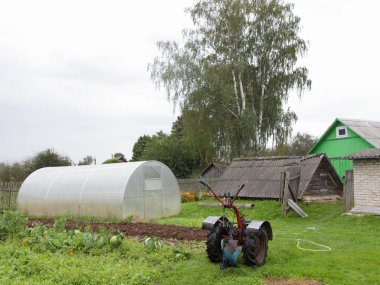 Kırsal tarım arazisi, kır arazisi - motor yetiştiricisi, sebzeli yataklar, eski bir ahırın arka planında modern polikarbonat serası ve yaz günü ağaçlar