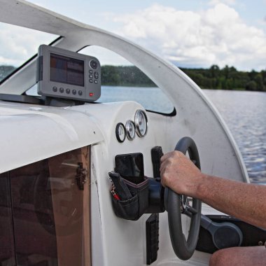 Kaptanın eli yüzen motorlu teknenin hidrolik direksiyonunda yakın çekim kabin kontrol paneli ve su ve nehir kıyısı arka planında yaz günü balık bulucu monitörlü kontrol cihazları.