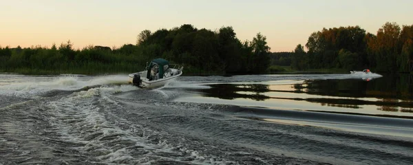 Fast gliding motor boats with turn water track on beautiful natural river landscape in calm water on forest background at dusk summer evening, watercraft rear view from water outdoor