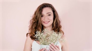 Portre. Kıvırcık kahverengi saçlı güzel bir kadın açık bir arkaplanda elinde çiçekler ve gülümsüyor.