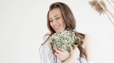 Güzel, mutlu, kahverengi saçlı, uzun saçlı ve makyajlı bir kızın portresi. Elinde çiçeklerle gülümseyen bir kız beyaz bir arka planda gülüyor.