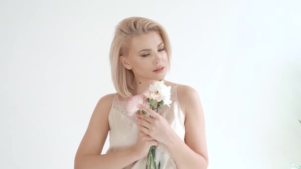 Портрет молодой привлекательной нежной блондинки в белом платье на белом фоне, которая держит букет белых цветов в руках, улыбается, флиртует, подмигивает — стоковое видео