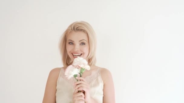 Retrato de uma jovem e atraente loira em um vestido branco sobre um fundo branco, que detém um buquê de flores brancas em suas mãos, mostra emoções — Vídeo de Stock