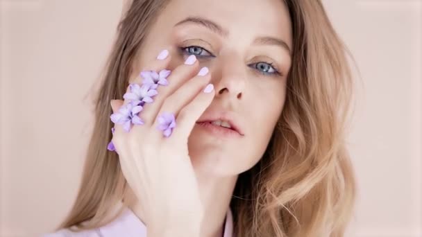 Portret młodej pięknej blondynki z długimi włosami i makijażem na jasnym tle, która biegnie ręką po twarzy, fioletowe kwiaty są wkładane między jej palce — Wideo stockowe