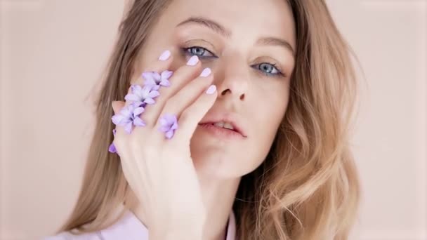 Retrato de una joven rubia hermosa con el pelo largo y maquillaje sobre un fondo claro, que pasa su mano sobre su cara, flores moradas se insertan entre sus dedos — Vídeos de Stock