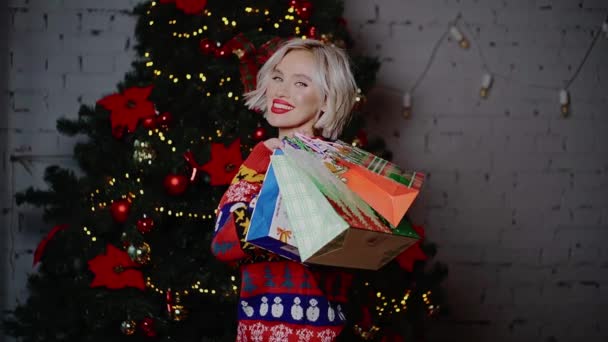 Young beautiful happy cute blonde with curdrs, red lipstick, gifts in her hands smiling, flirts, buys gifts near the Christmas tree indoors — Stock Video