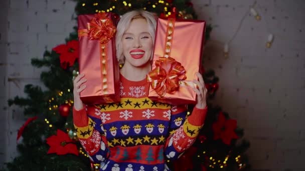 A young cute happy, blonde woman with blue eyes and white skin holds a red gift box near the Christmas tree indoors — Stock Video