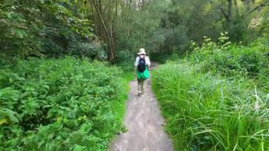 Genç bir kız yaz ormanında ağaçlar ve çimenler arasında orman yolunda yürüyor..