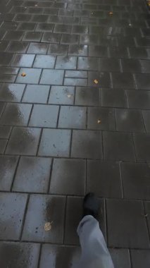 Kotlu ve spor ayakkabılı bir adam yağmurda ıslak sokak fayansları üzerinde yürür. Dikey video.