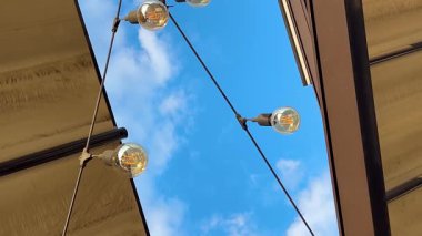 Açık hava çelenkleri bir restoranda dizilmiş dekoratif ampullerle mavi gökyüzü ve bulutların arkaplanına karşı..