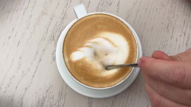 Süt köpüğünde tüy ya da yaprak şeklinde latte sanatı olan bir fincan latte. Güzel bir kahve tasarımının yakın çekimi. Bir kaşık süt köpüğüyle kahve karıştırır..