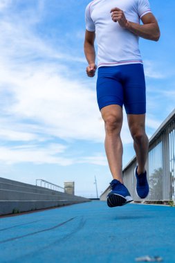 Mavi pantolonlu ve beyaz tişörtlü genç koşucu mavi bir pistte güneşte koşuyor. Motivasyon konsepti, kariyer