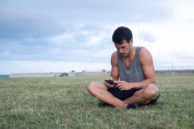 Kafkasyalı genç adam çimenlerde gri bir gömlek giyerek otururken akıllı telefonuna bakıyor.