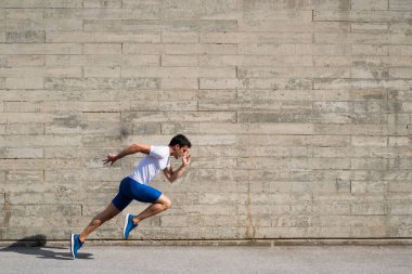 Sporcu, düzensiz gri duvar arkaplanının önünde mavi pantolon ve spor ayakkabı giymeye başlıyor.