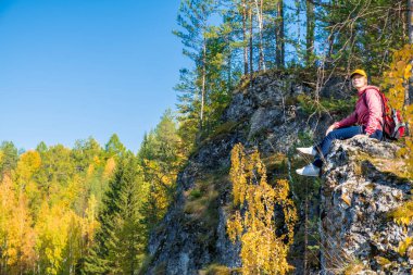 Sırt çantalı kız turist, Rusya 'nın Ural kentinde bir kayanın üzerinde otururken sonbahar ormanına bakıyor..