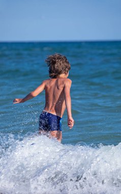 Sırtı olan mutlu bir çocuk suya daldırma ve dalgalarla yürür, dikey biçimde.
