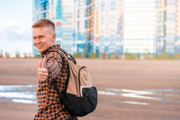 A happy young man in a checked shirt with a backpack gives a thumbs-up against the background of multicolored multi-storey new buildings, first real estate purchase, affordable housing for young family