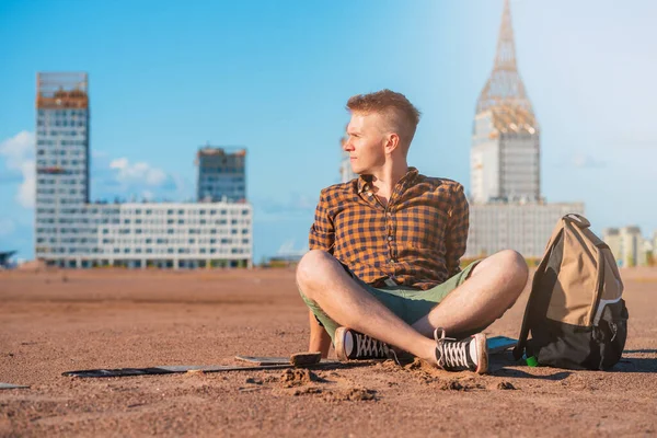 A young man in a checked shirt with a backpack sits on a sandy wasteland, behind multi-storey buildings under construction, waiting to buy his own apartment