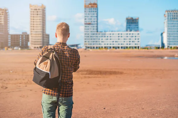 young man in a checked shirt with a backpack stands on a vacant lot in front of multi-storey buildings under construction, waiting for settlement