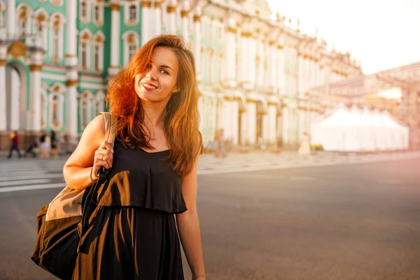 A young brunette woman in a dress with long hair with a backpack smiles against the background of the winter Palace on the Palace square in St. Petersburg