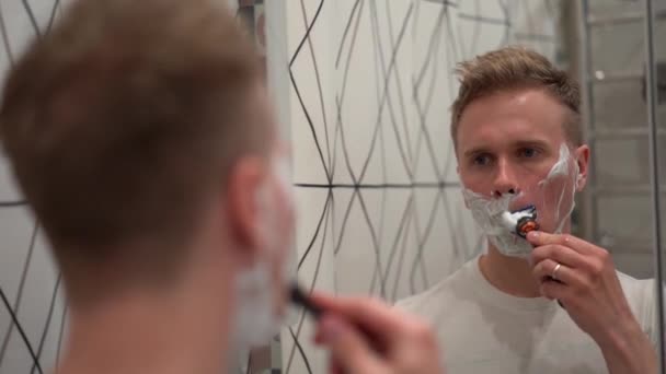 白いバスルームにシェービングフォームを適用した後ブロンドの男は彼の頑丈を剃ります — ストック動画