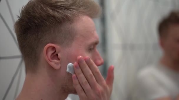 一个金发碧眼的男人在一个白色的浴室里用剃须泡沫刮掉了他的胡子 — 图库视频影像