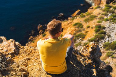 Sarı elbiseli genç adam denizin üzerinde oturuyor, kıyı kayalarının arkasında, sakin açık mavi deniz, Kırım 'da Cape Fiolent.