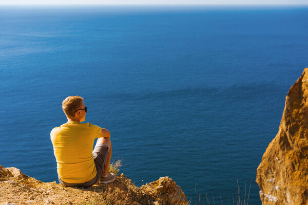 Young man in yellow clothes sitting high above the sea, against the background of coastal rocks, calm clear blue sea, Cape Fiolent in Crimea.
