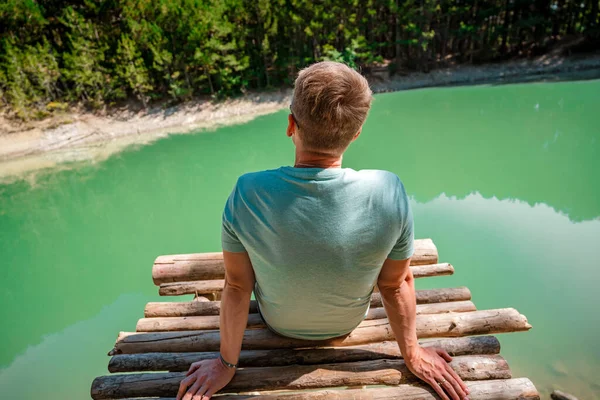 Young blond man on a bridge above a mountain lake with clear water and a view of a green forest. Copy space