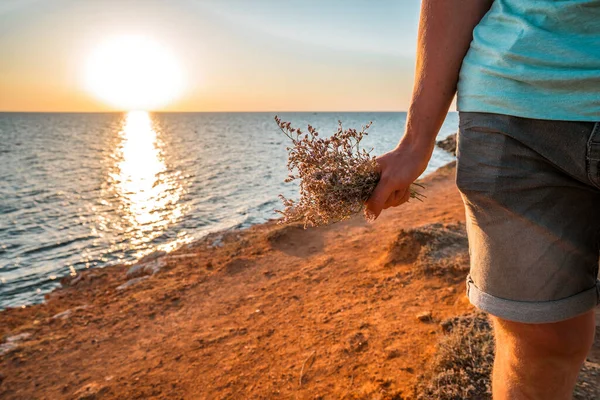 一个男人站在岩石上俯瞰美丽的日落海景时 手里拿着一束鲜花 — 图库照片