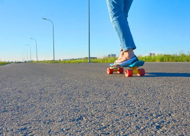 Mavi kotlu ve parlak renkli spor ayakkabılı bir kız güneşli bir günde asfalt bir yolda mavi bir kaykaya biniyor..                    