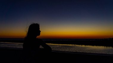 Açık mavi turuncu gün batımı denizlerin üstünde ve sırtından genç bir kadının silueti.