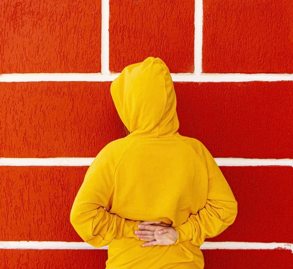 Teenager girl in a yellow sweatshirt or hoodie in a hood against the background of a red wall with white seams