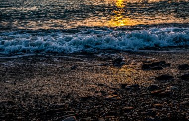Gün batımında deniz manzarası. Deniz suyunda güneşli bir patika ve çakıl taşı sahilinde yaklaşan bir dalga. Manzara
