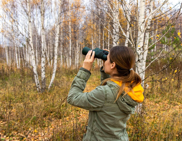 Young woman birdwatcher with binoculars in the autumn forest. Birdwatching, zoology, ecology. Research, observation of animals. Ornithology