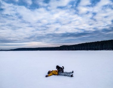 Sarı ceketli genç bir kadın karlı bir gölde gri tüylü bir köpekle oynuyor. Hayvanlarla yürüyorum. Cep telefonu fotoğrafı. Kız köpeği gezdirir. Kış manzarası