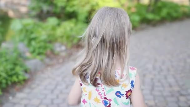 Ένα μικρό ξανθό κορίτσι περπατά σε ένα πέτρινο δρόμο ένα πάρκο ή ένα δάσος. Φόρεμα λουλουδιών. — Αρχείο Βίντεο